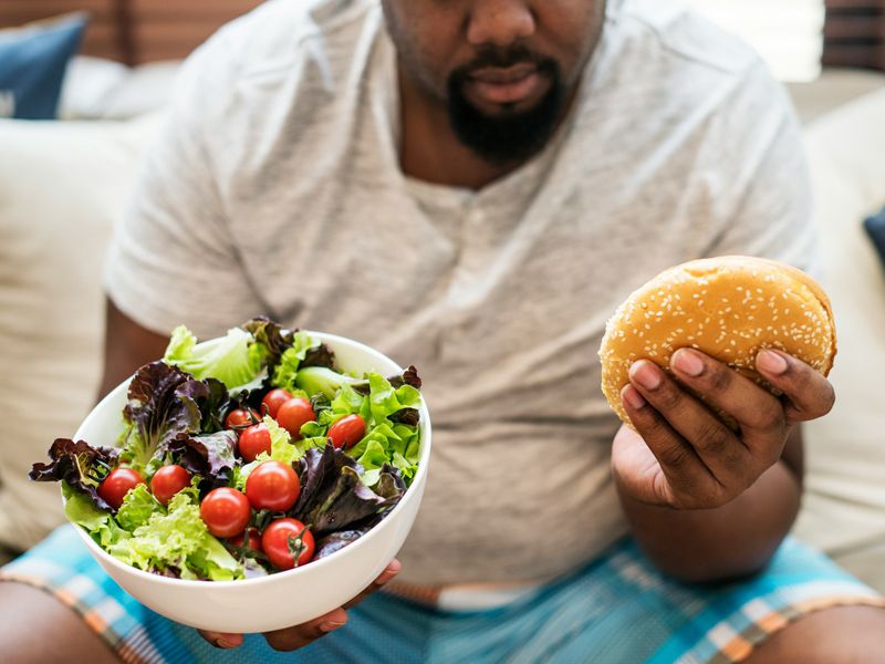 Man holding a bowl of salad in one hand and a burger in the other, symbolising mindful eating, healthy choices, and the balance needed for lasting weight loss - Retrained Minds Hypnotherapy.