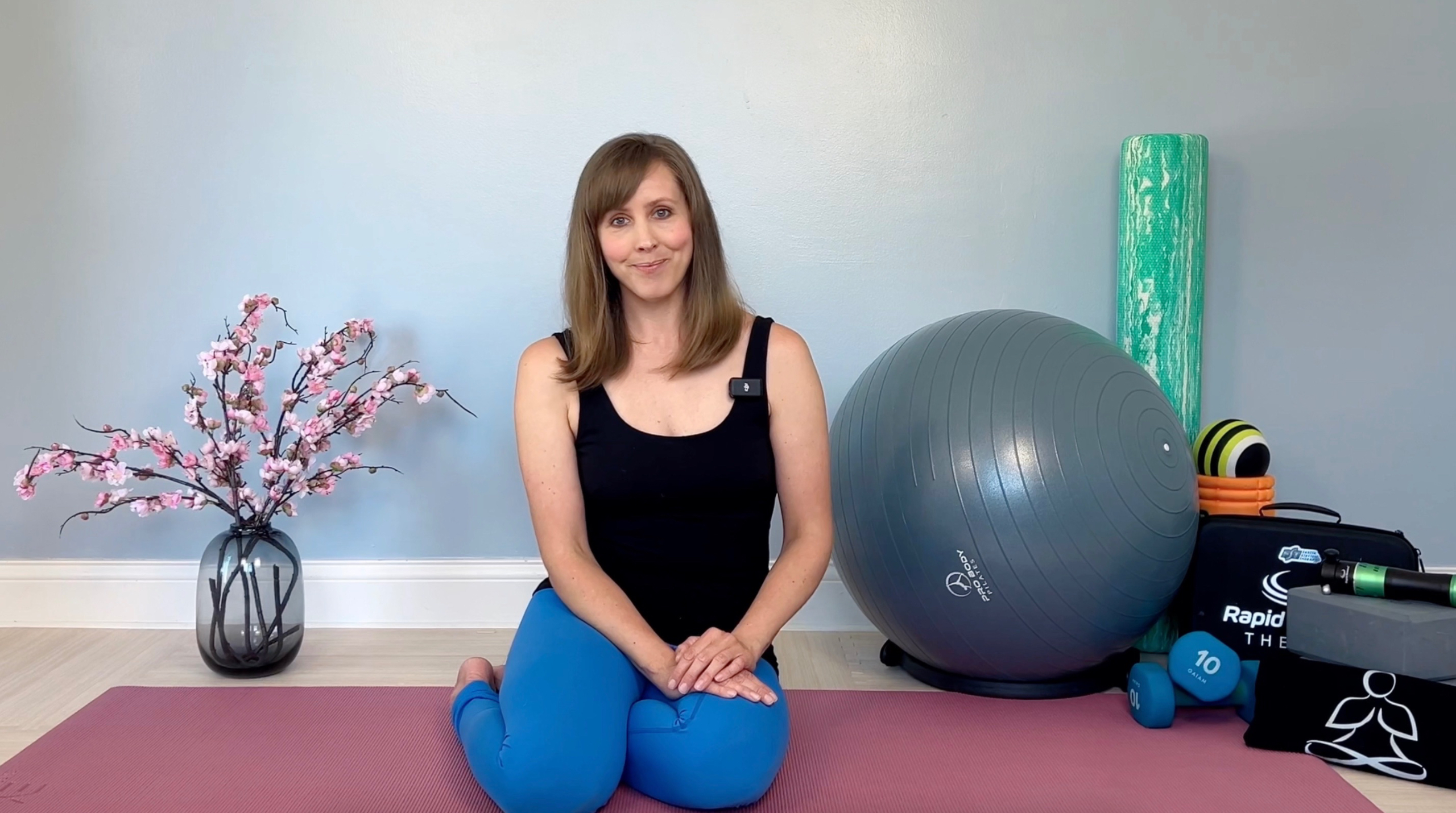 Shannon Hershman of Be Well My Body—On Demand discusses classes & courses while on a pink yoga mat with a ball and fitness equipment behind her.