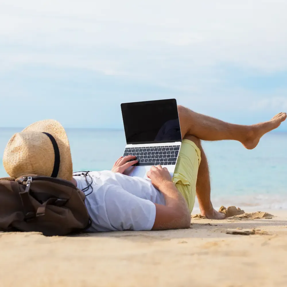 A man relaxing on a sandy beach works on a laptop while lying down with one leg crossed. He’s wearing a straw hat, white shirt, and yellow shorts, with a brown backpack beside him. The ocean and clear blue sky create a peaceful backdrop.