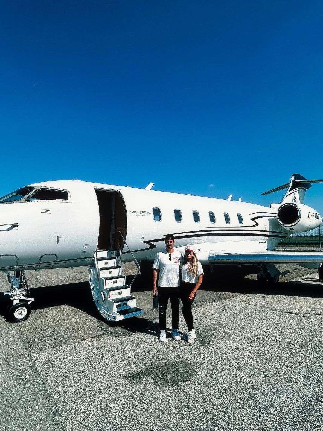 A man and woman stand together on an airport tarmac in front of a white private jet with the words “DARE TO DREAM” near the cabin door. Both are dressed casually in white tops and black pants under a clear blue sky, with the jet’s stairs extended beside them.