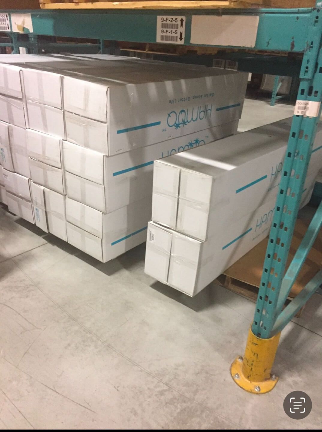 Stacks of white rectangular boxes labeled “Hammoq” stored on the floor beneath a metal warehouse shelving unit. The boxes are sealed and organized neatly, with visible blue branding and lines on the packaging.