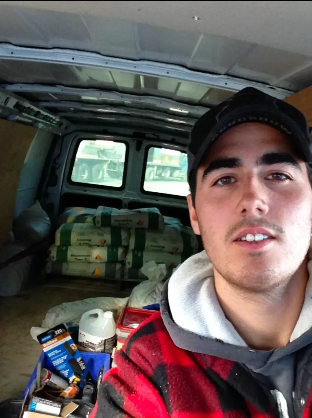 A man wearing a black cap, gray hoodie, and red plaid jacket takes a selfie inside a work van. Behind him are stacked bags of construction materials and various tools and supplies, including bottles, boxes, and containers. The van’s interior is metallic with wooden panels on the sides.