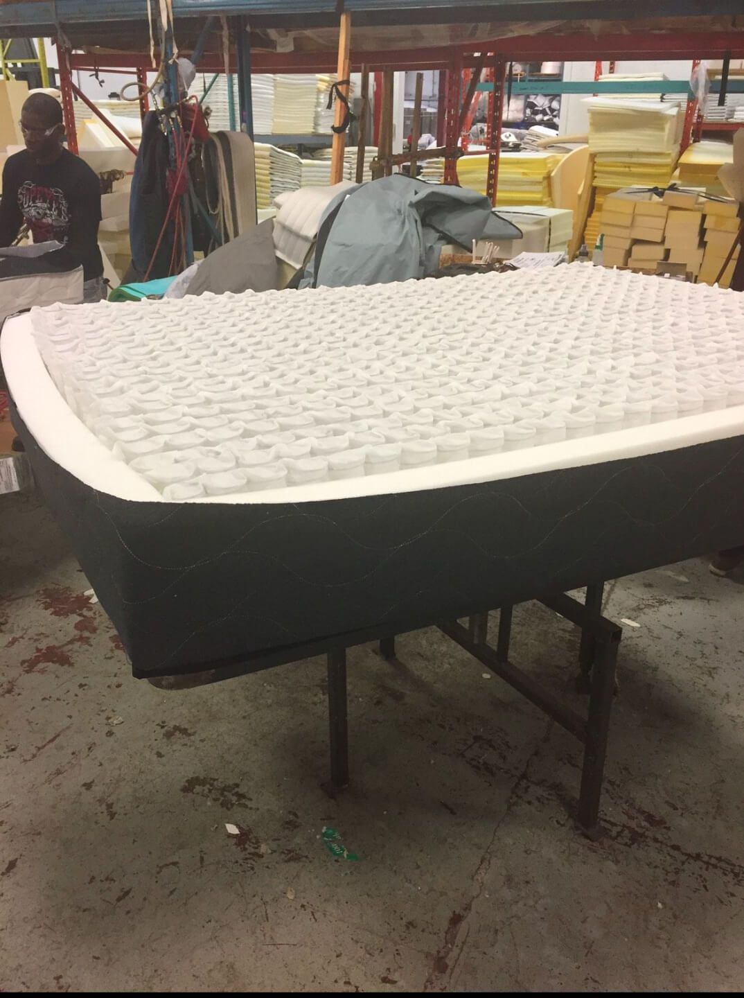 A partially assembled mattress on a metal worktable in a factory setting. The mattress has visible foam layers and individually wrapped coils. In the background, stacks of foam and other bedding materials are visible, along with a worker wearing a dark shirt and safety glasses.