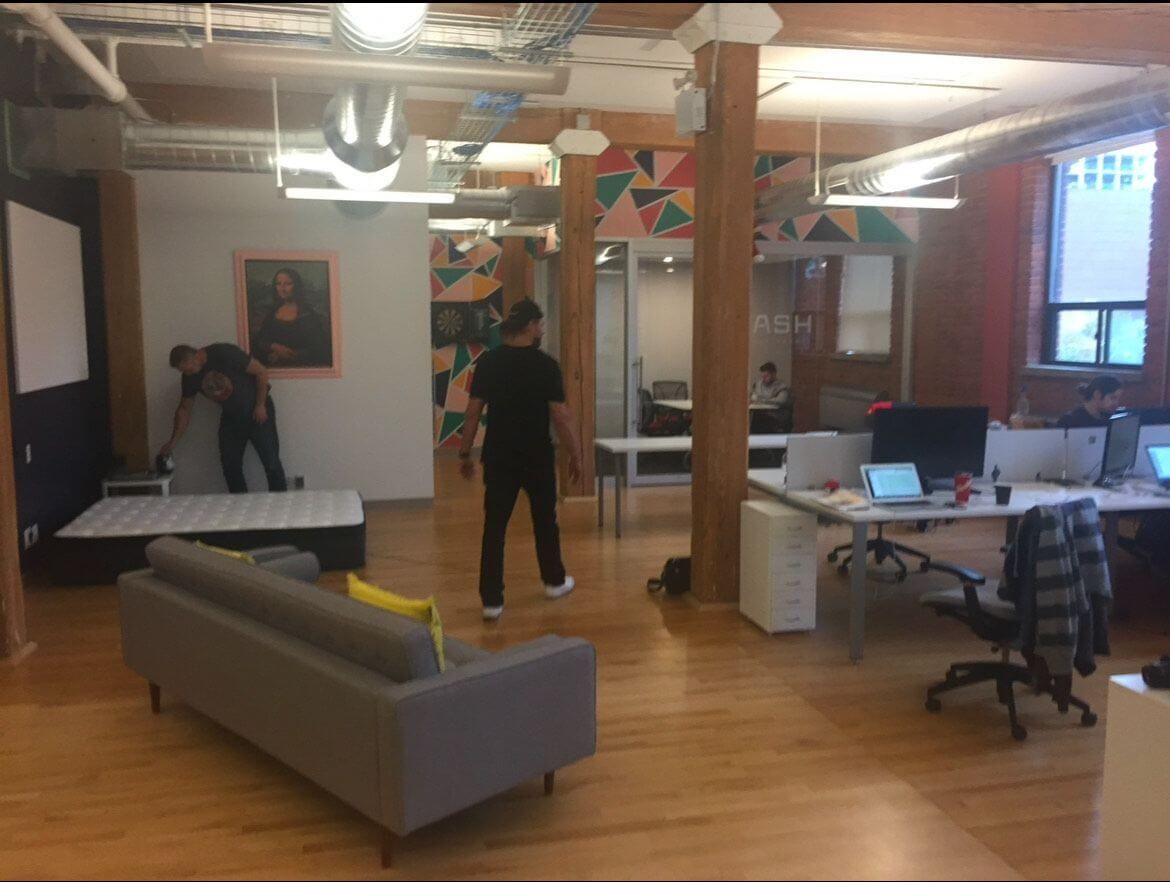 A modern open-concept office with wooden floors and exposed beams. Two men are setting up a mattress in one corner near a framed Mona Lisa print, while others work at desks with laptops and monitors. The room features colorful geometric wall art, industrial air ducts, and natural light from large windows.
