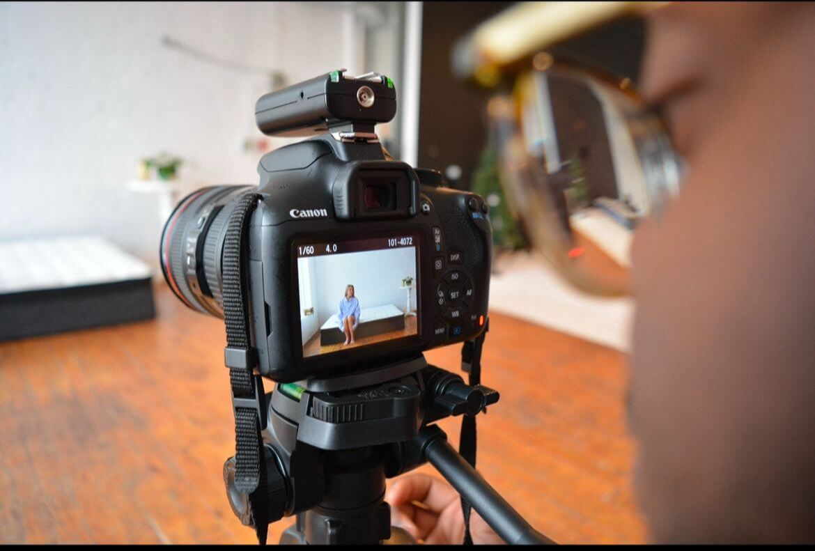 A photographer is capturing a model sitting on a bed in a bright studio. The focus is on the Canon camera’s screen, showing the model seated at the center of the frame, while the photographer’s face and glasses are blurred in the foreground. The studio has wooden floors and a minimalist setup.