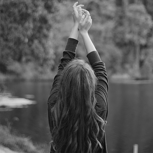 woman looking out at a lake holding her arms up in celebration of life