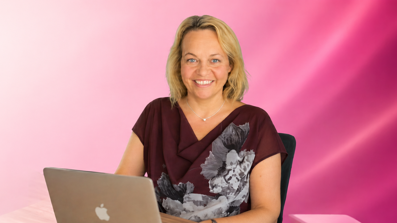 Coach Aga - a smiling white women, blonde with hair to her shoulders and purple top, sitting at her laptop.