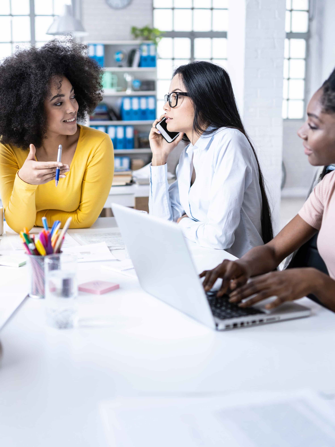 “Three women working together in an office, with one speaking on the phone and another using a laptop.”