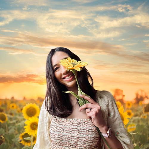 image of woman holding a sunflower that covers part of her face. She is standing in a field of sunflowers.