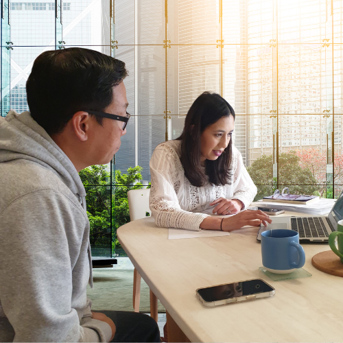office environment. A women is showing her laptop to a man. They are both seated at a desk. 