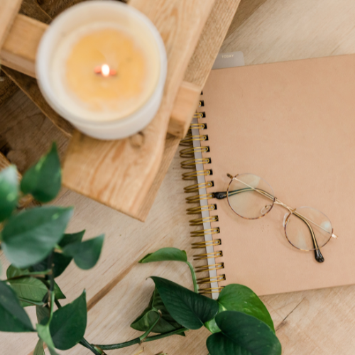 image of a notebook, glasses, candle and plant sitting on a wooden desk