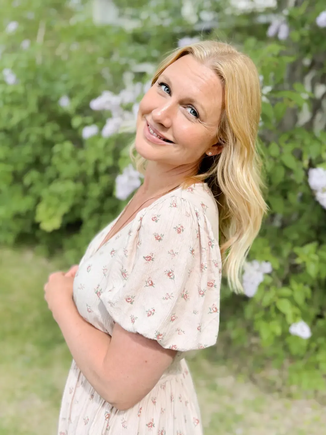 A woman smiling and looking at us. Standing by a hedge with flowers, wearing a floral summer dress.