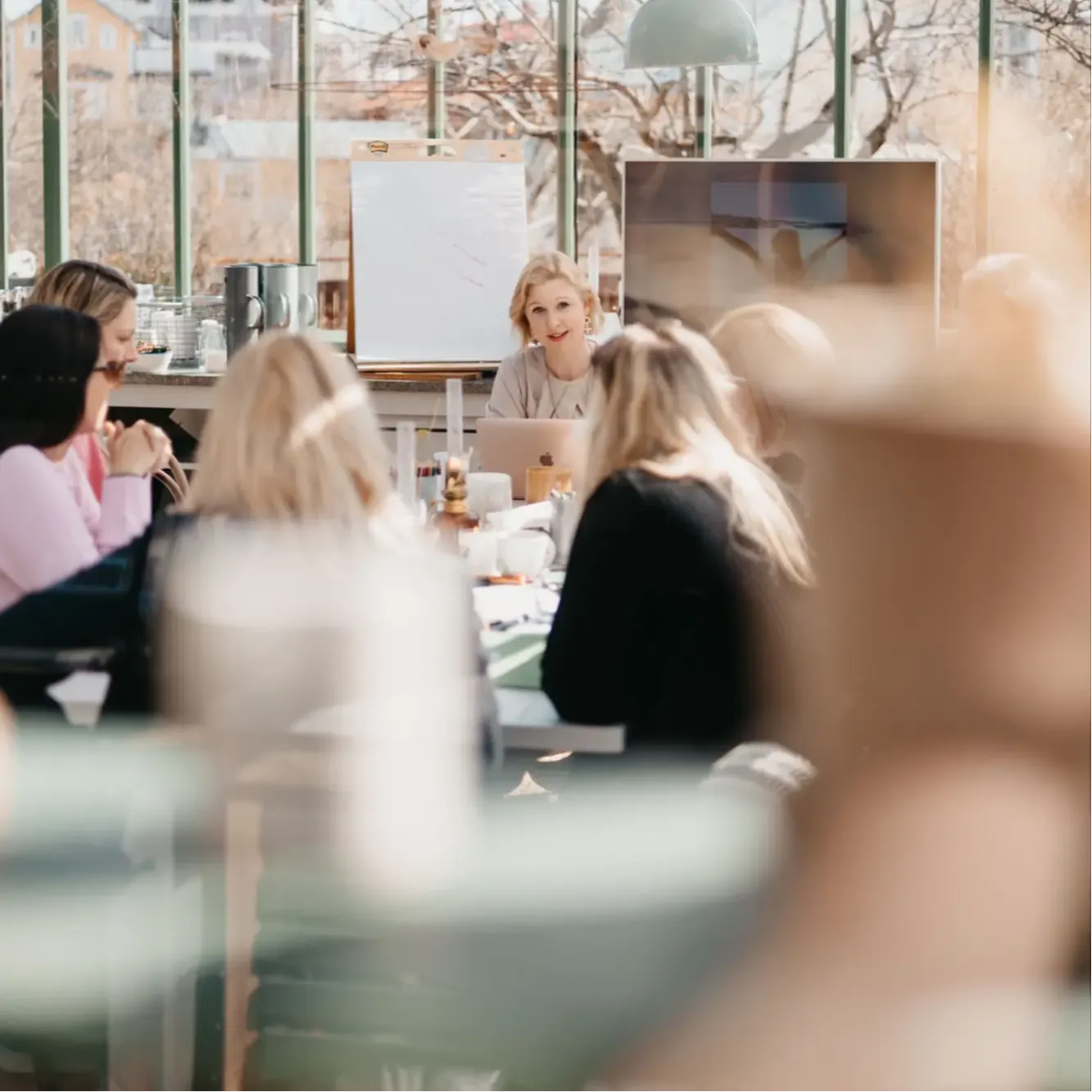 A room full of women sitting by a table. The woman at the head of the table is speaking to the other women who's listening intently.