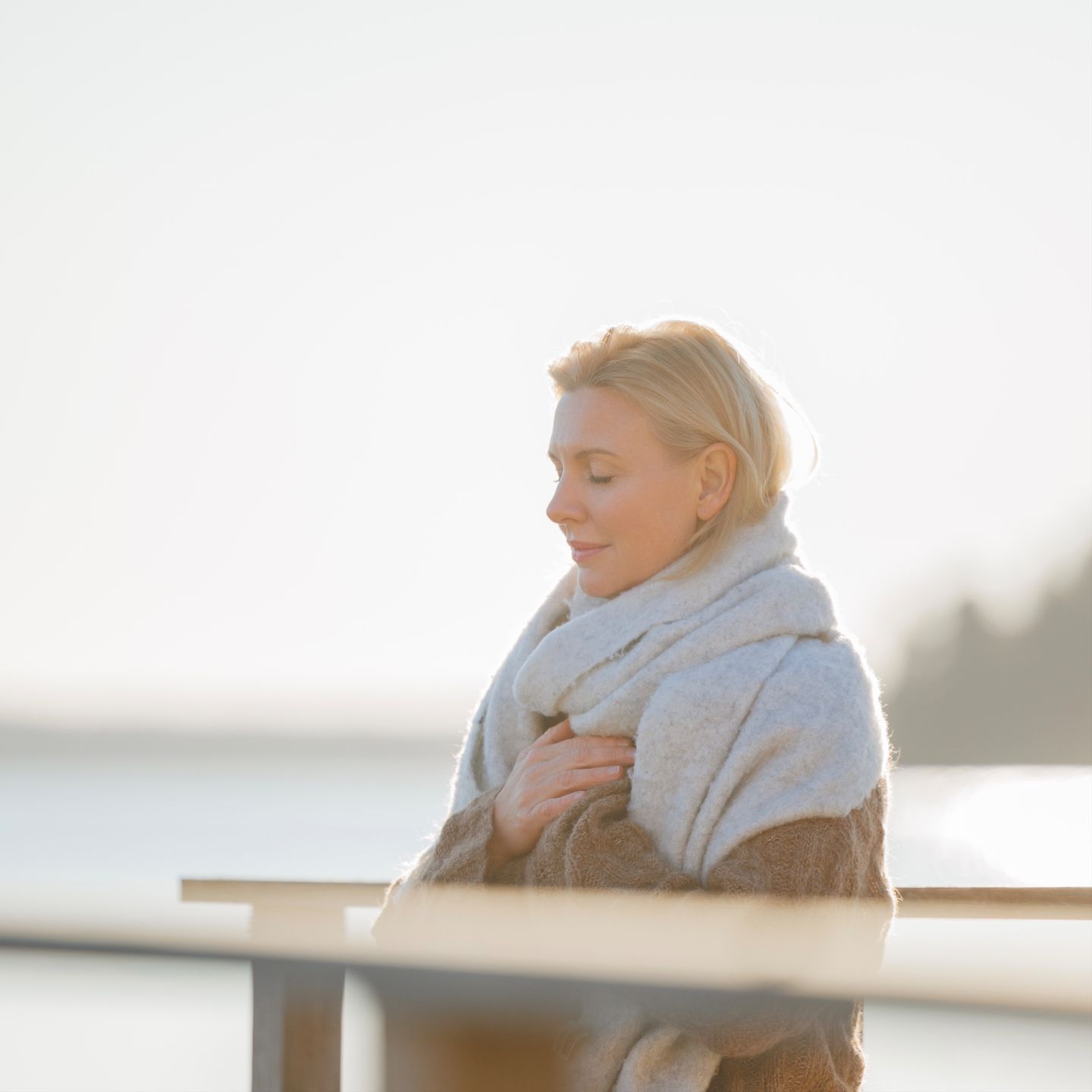 Woman is standing outside, turning around swaying her arms and looking down in the ground with a smile. In the distance you can see a lake.