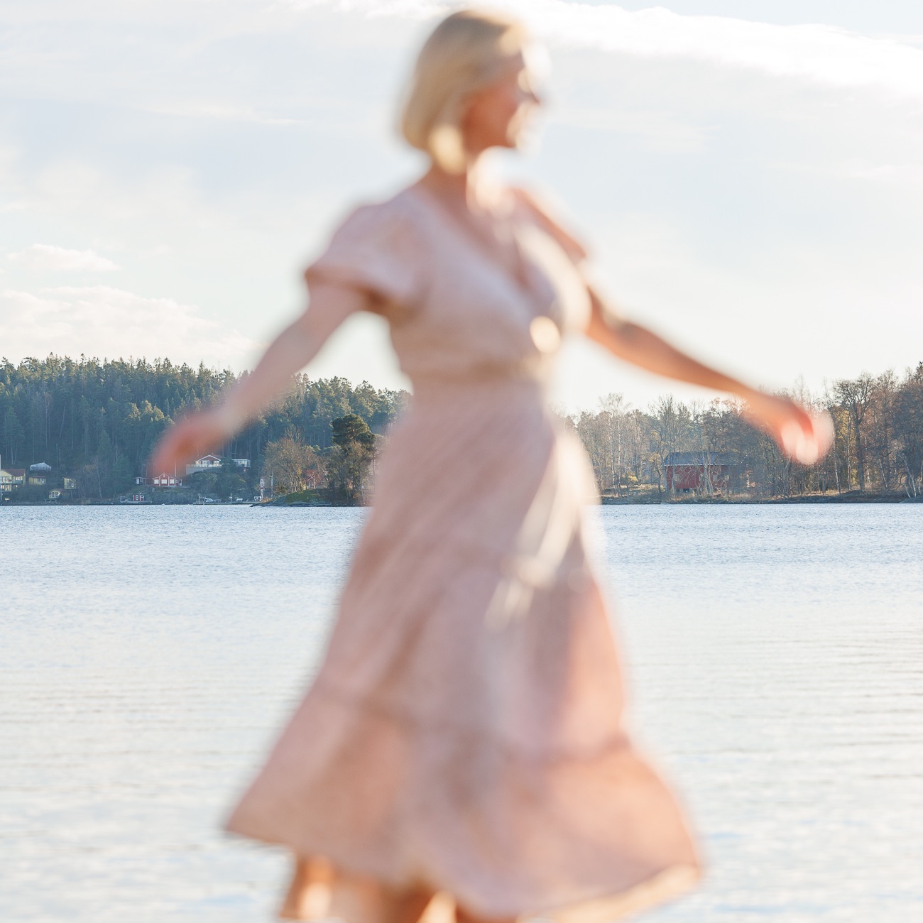A woman smiling and looking at us. Standing by a hedge with flowers, wearing a floral summer dress. 