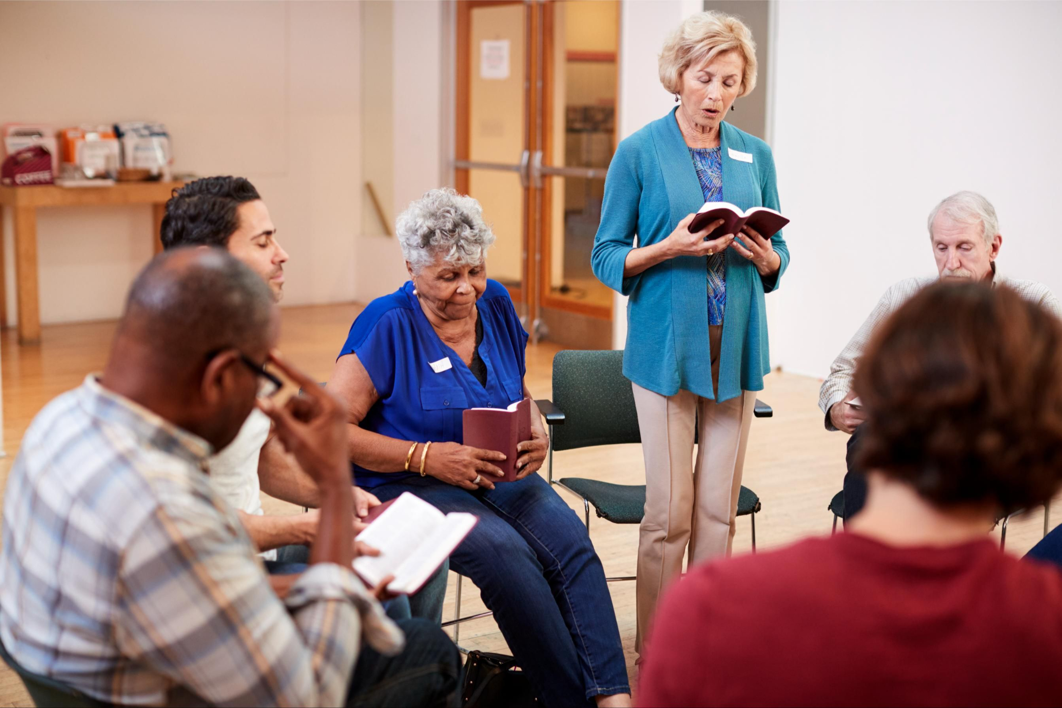 Group of older people in a circle, one is women is standing and reading from a book.