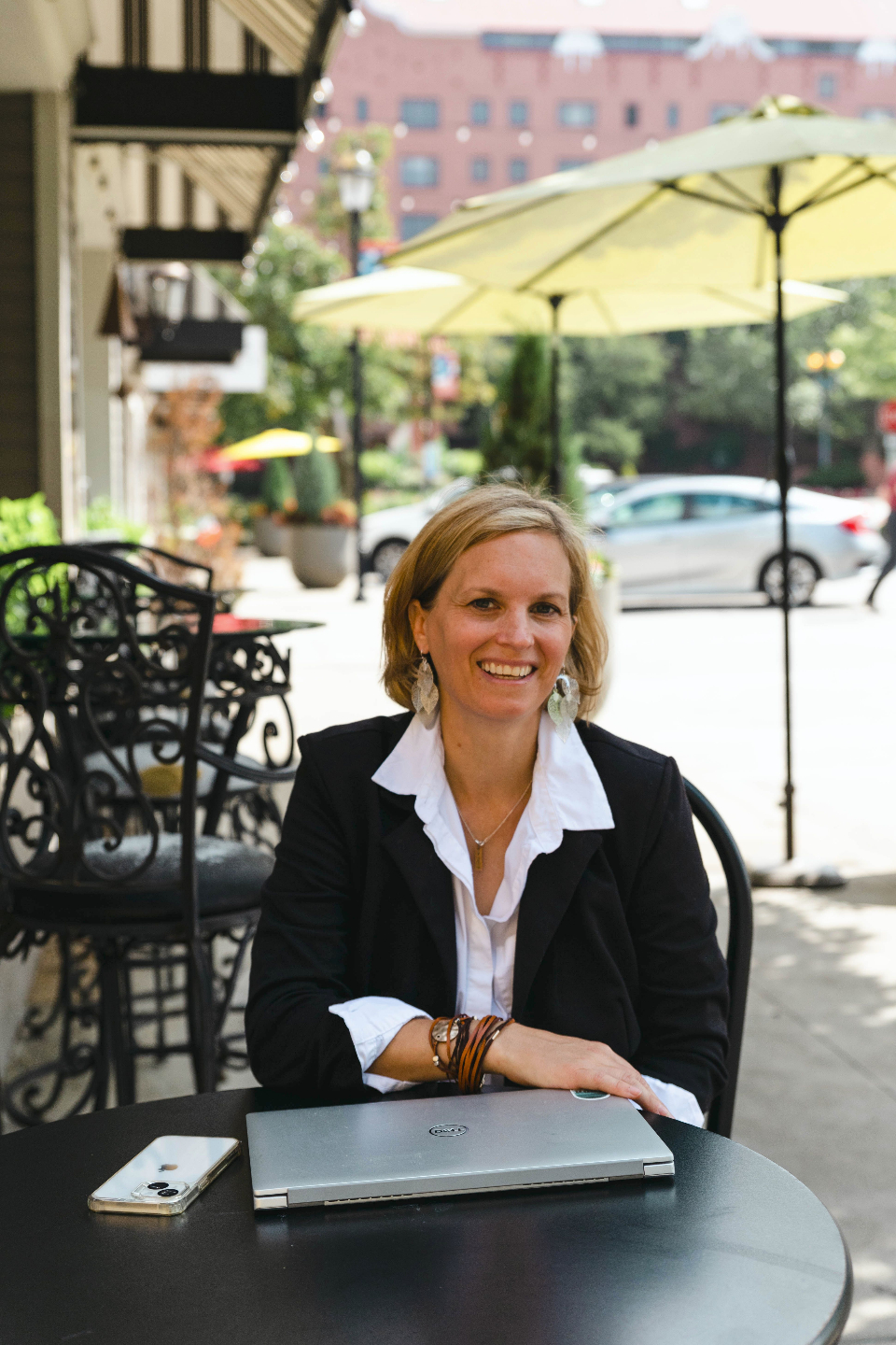 Andrea Anderson is sitting at an outside cafe table and is smiling, wearing a black blazer and white shirt. There is a closed laptop and white iphone on the table. In the background are yellow umbrellas and cars 