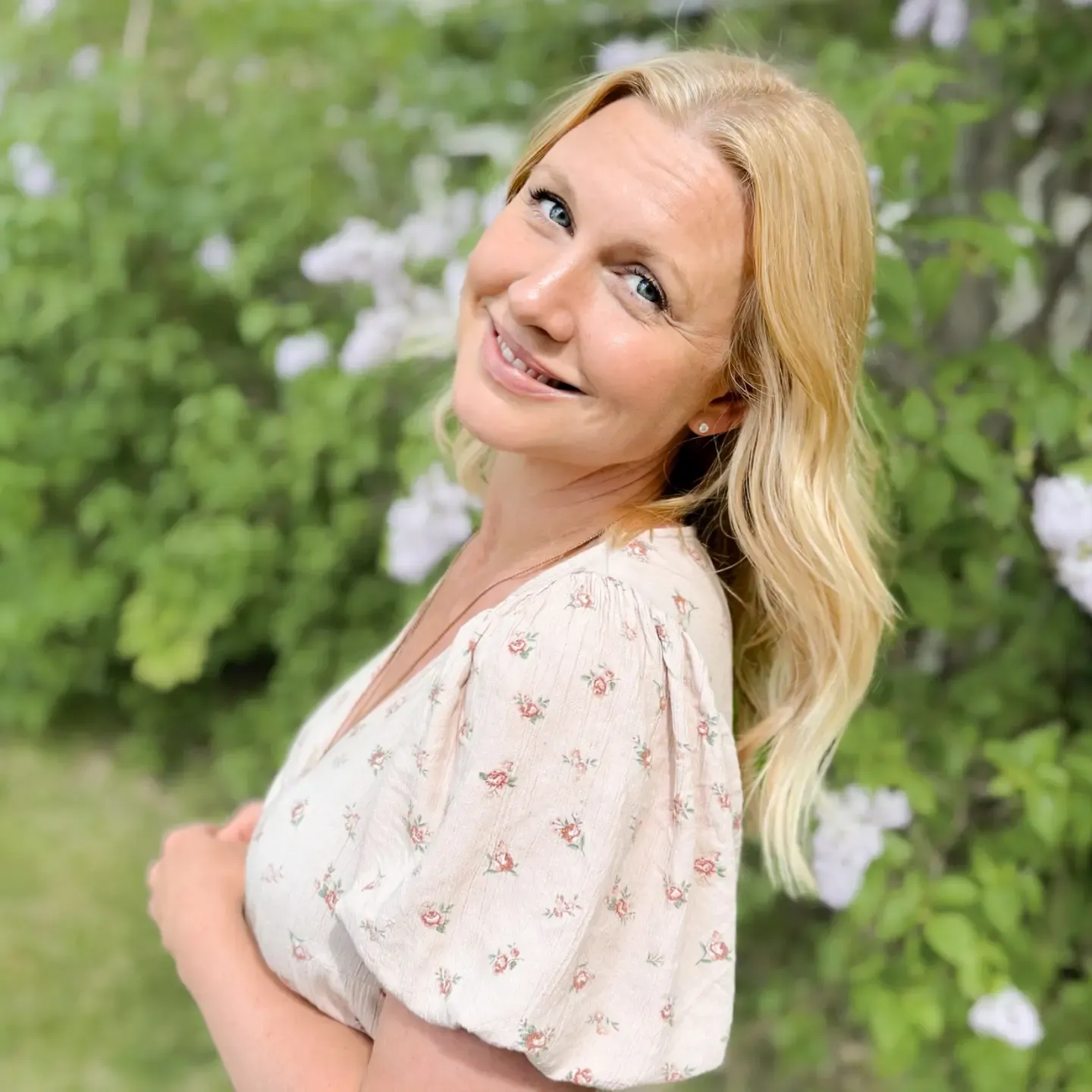 Sofia looking into at us. She is standing outside by a hedge with flowers. She is smiling and wearing a floral summer dress.