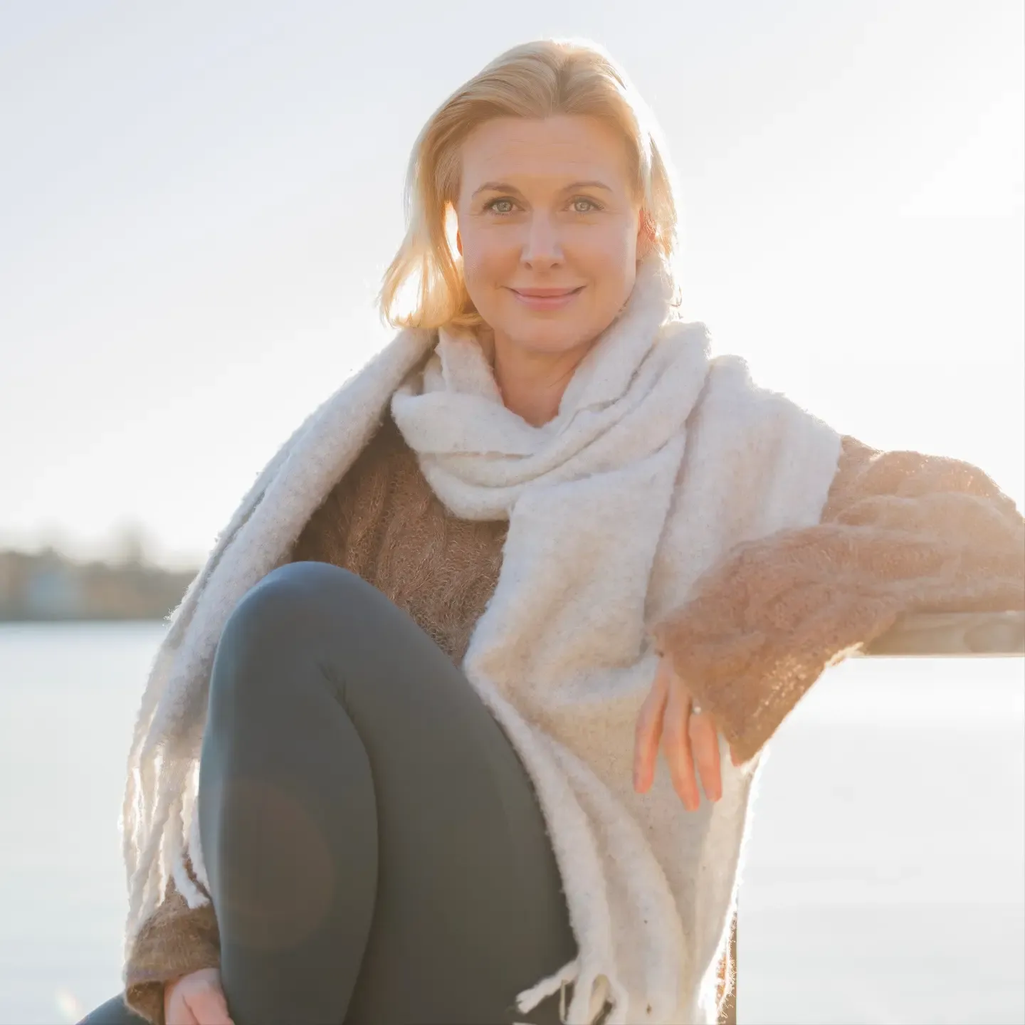Sofia looking into at us. She is standing outside by a hedge with flowers. She is smiling and wearing a floral summer dress. 