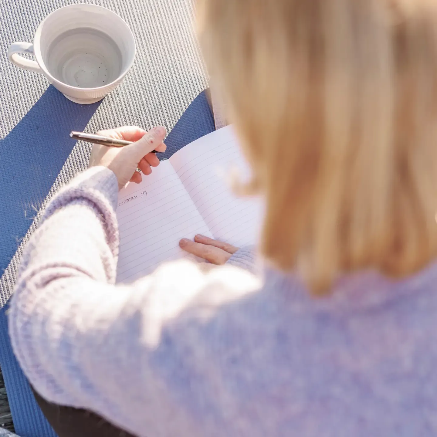 Woman writing in a book, there is a cup on the table. The sun is shining on the woman.