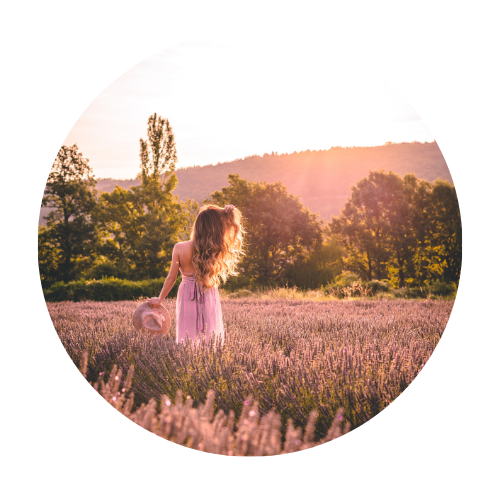 woman, walking away. She is in a field of lavender flowers, holdiing a hat. 