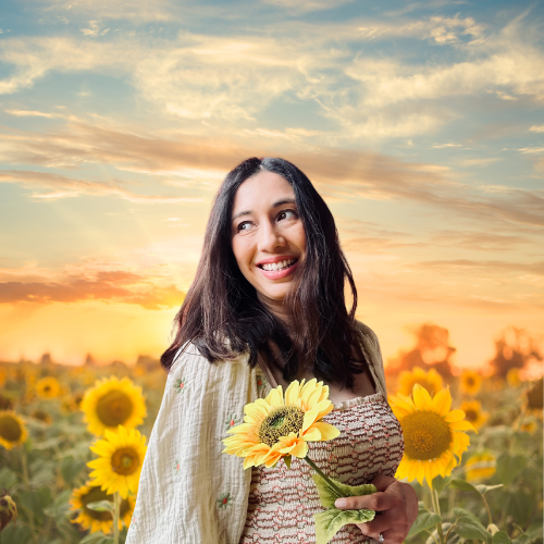 woman smiling, holding a sunflower. She is standing in a field of sunflowers with the sun behind her. 