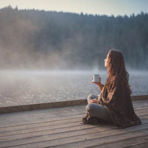 women sitting cross legged on a wooden surface. She is holding a cup and looking out across the water.