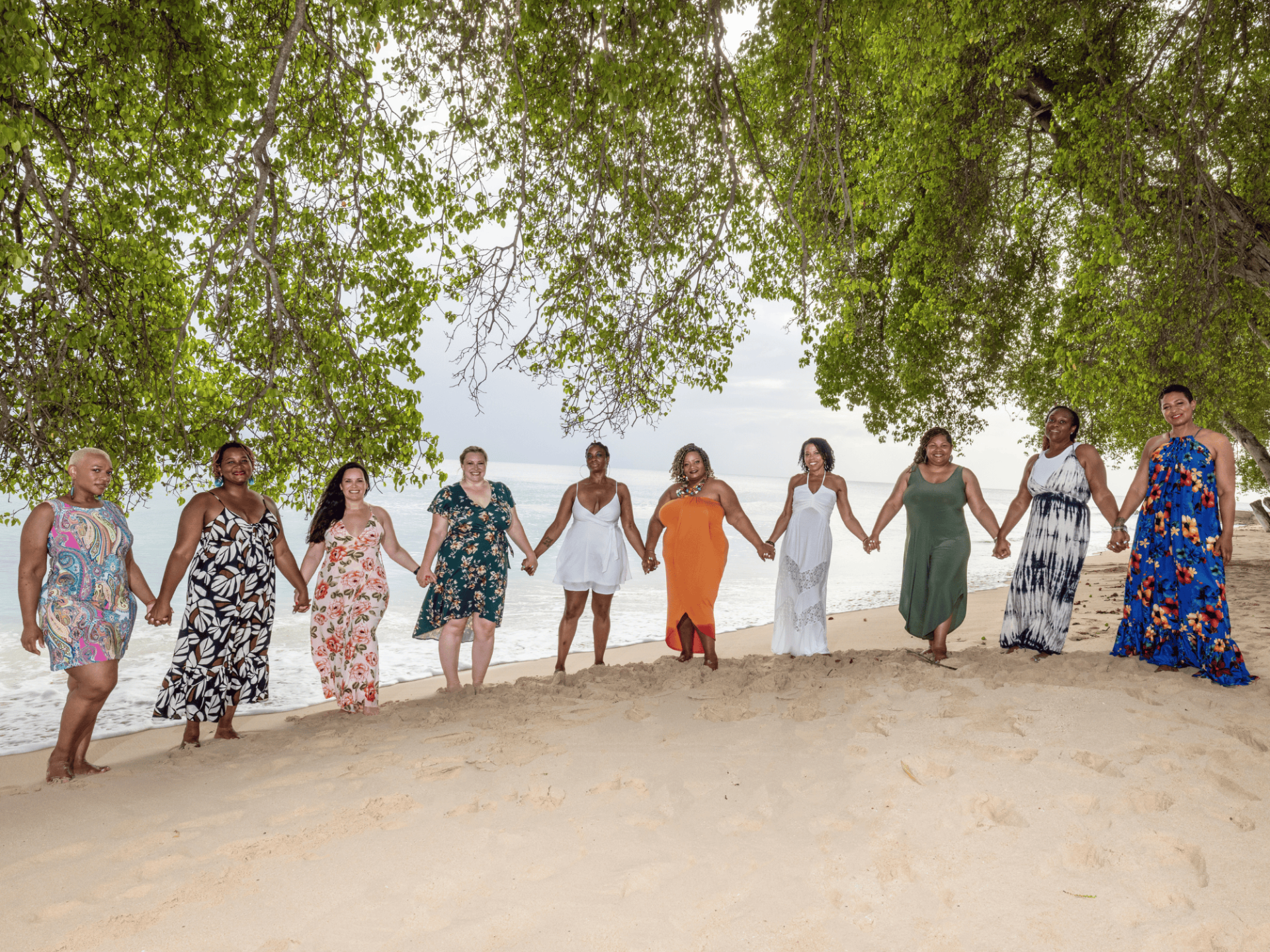 10 women smiling warmly and holding hands on the beach in colorful sundresses, facing the camera. Jamila in orange in the center with 9 multi ethnic women in front of the sea. Tree foliage is above them and light sand is in front of them