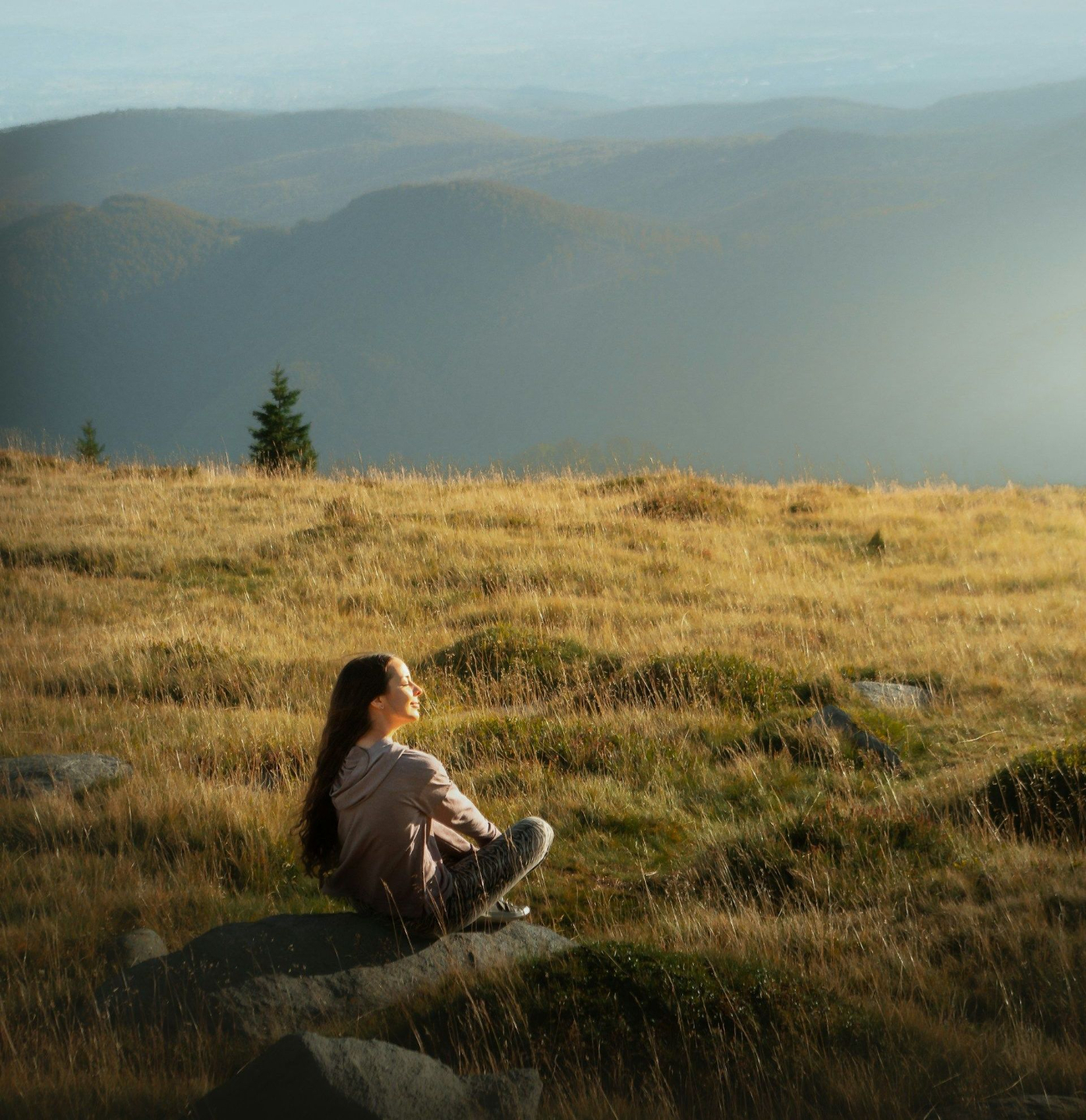 woman sitting on a large stone in a grass field on the top of a mountain overlooking a mountainscape