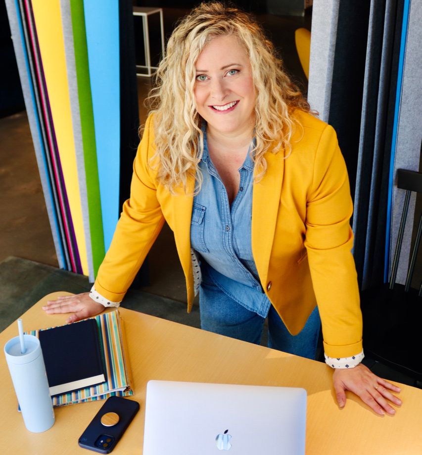 Photo of Stephanie Broquist in a yellow blazer standing at a desk and looking into the camera smiling.