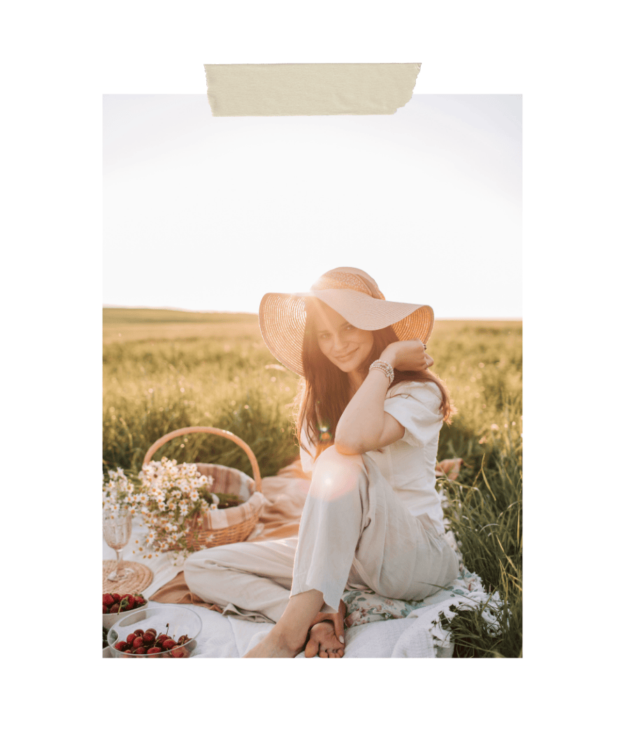 Woman sitting on a picnic blanket in a sunny field wearing a wide-brimmed hat, surrounded by flowers, fruit, and a wicker basket, representing calm, mindfulness, and outdoor relaxation.