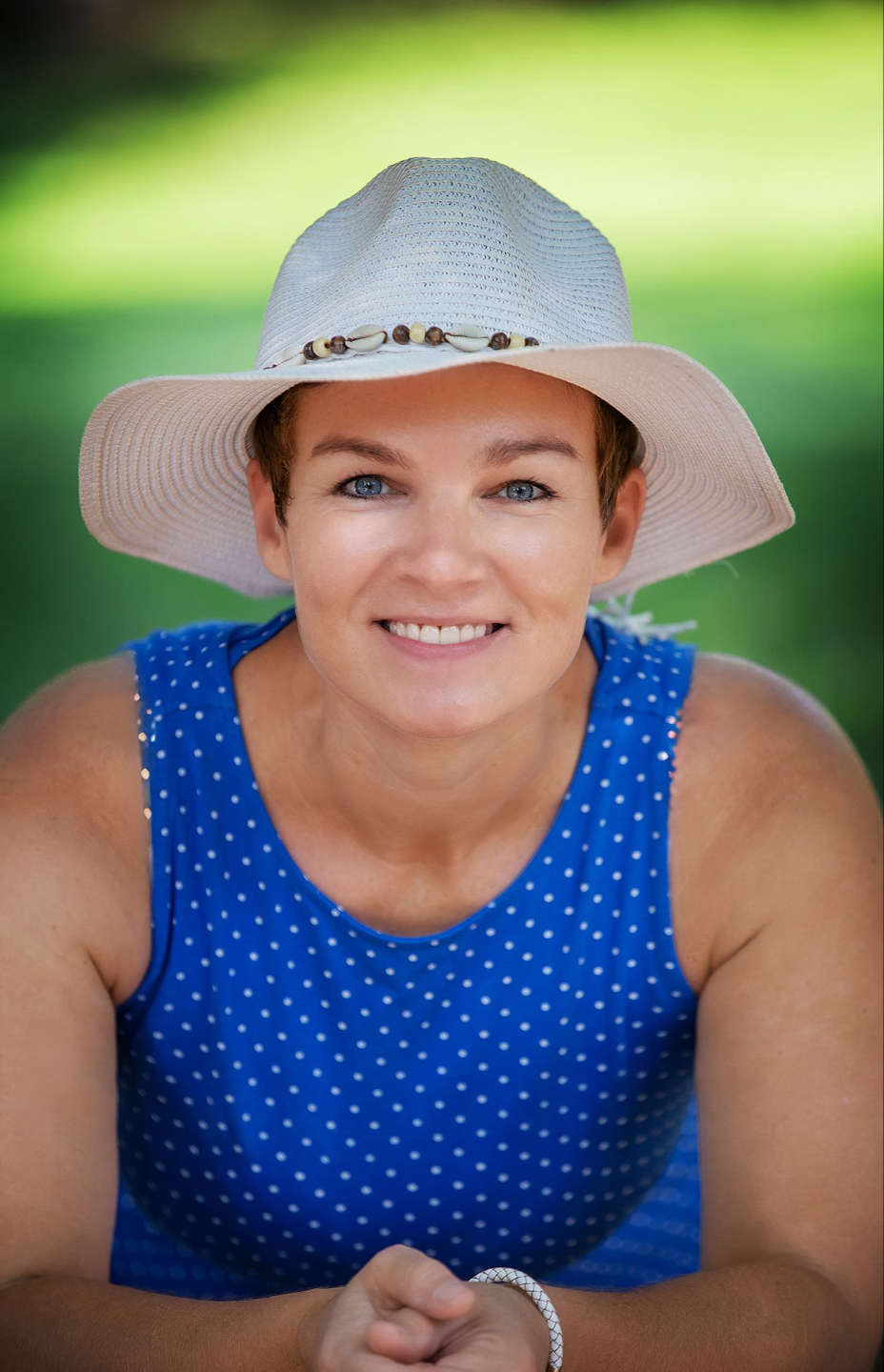 Kathi trägt ein weißes Kleid und einen weißen Hut, hockt hinter einem grünen Gebüsch und lächelt sanft – eine Szene, die Ruhe, Natürlichkeit und einen Hauch von Verspieltheit ausstrahlt.