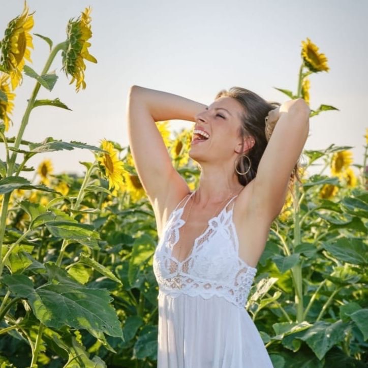 Somatic healing practitioner Emily Wehrman smiling with her hands by her head in a field of sunflowers.