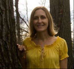 Natalie Gensits, a smiling woman with shoulder length light brown hair wearing a yellow top and leaning against a tree