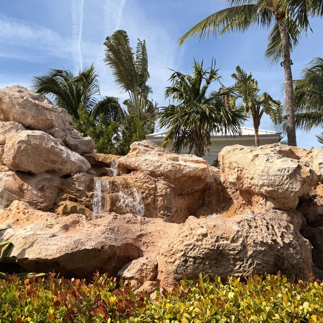 Baha Bay at Baha Mar: Rock waterfall feature at Baha Bay, framed by palm trees and tropical plants under a sunny blue sky.