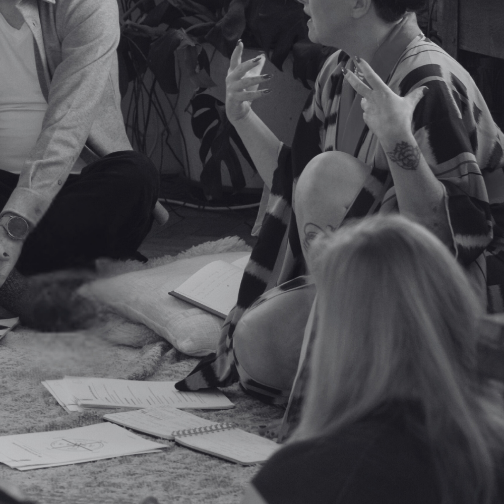 Black-and-white photo of a group of participants sitting in a circle during a Female Domination workshop, focused on conscious scene creation and power dynamics.
