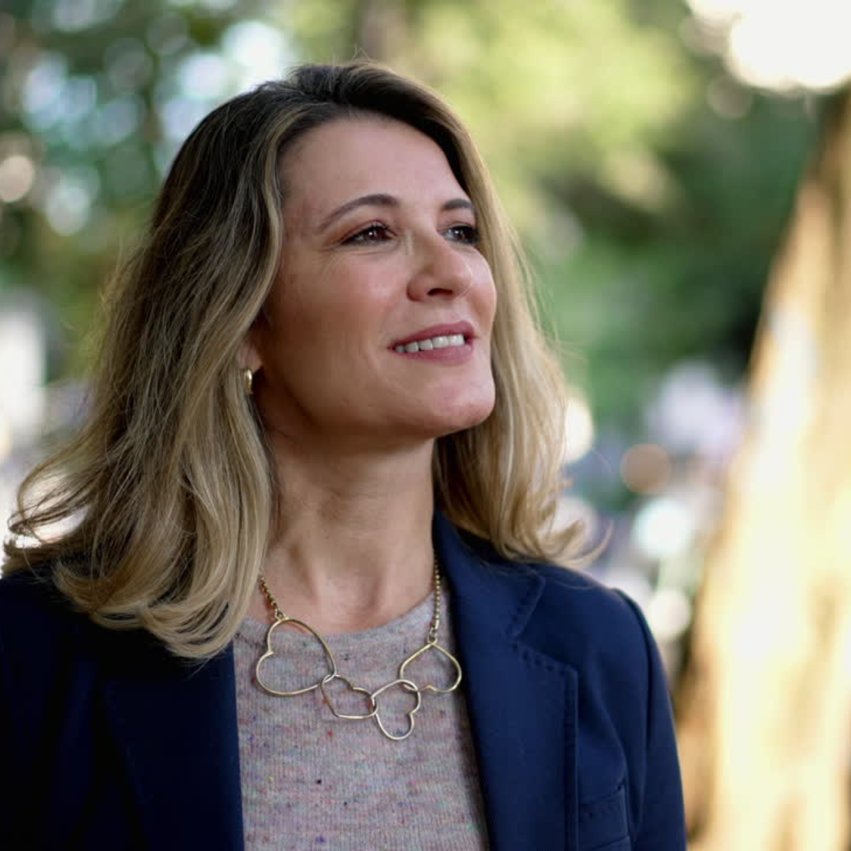 Image of woman with blond hair, black blazer, and tan shirt standing outside in nature