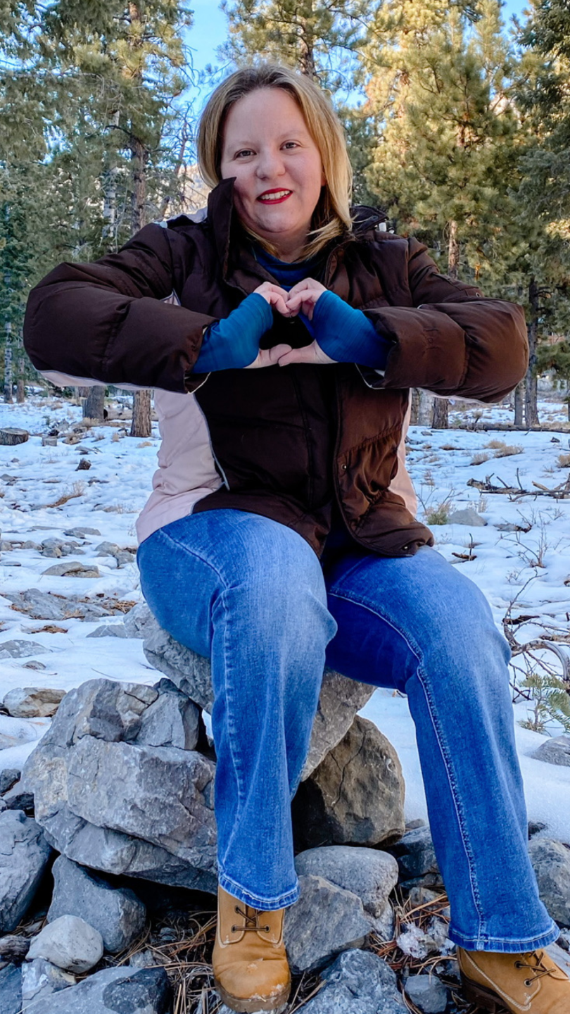 Image of Melinda in the mountains with a brown and pink jacket with blue jeans and brown boots. Snow on the ground and pine trees behind her.