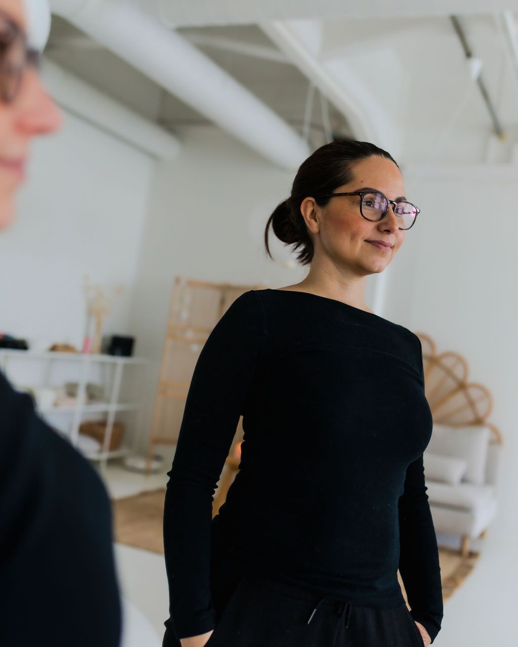 Susanna istuu puisen pöydän ääressä nojaten kyynärpäitään pöytään ja katsoo hymyillen kameraan. Hänellä on musta pitkähihainen paita ja mustasankaiset silmälasit. Tausalla näkyy boho chic tyylinen rauhallinen huone.
