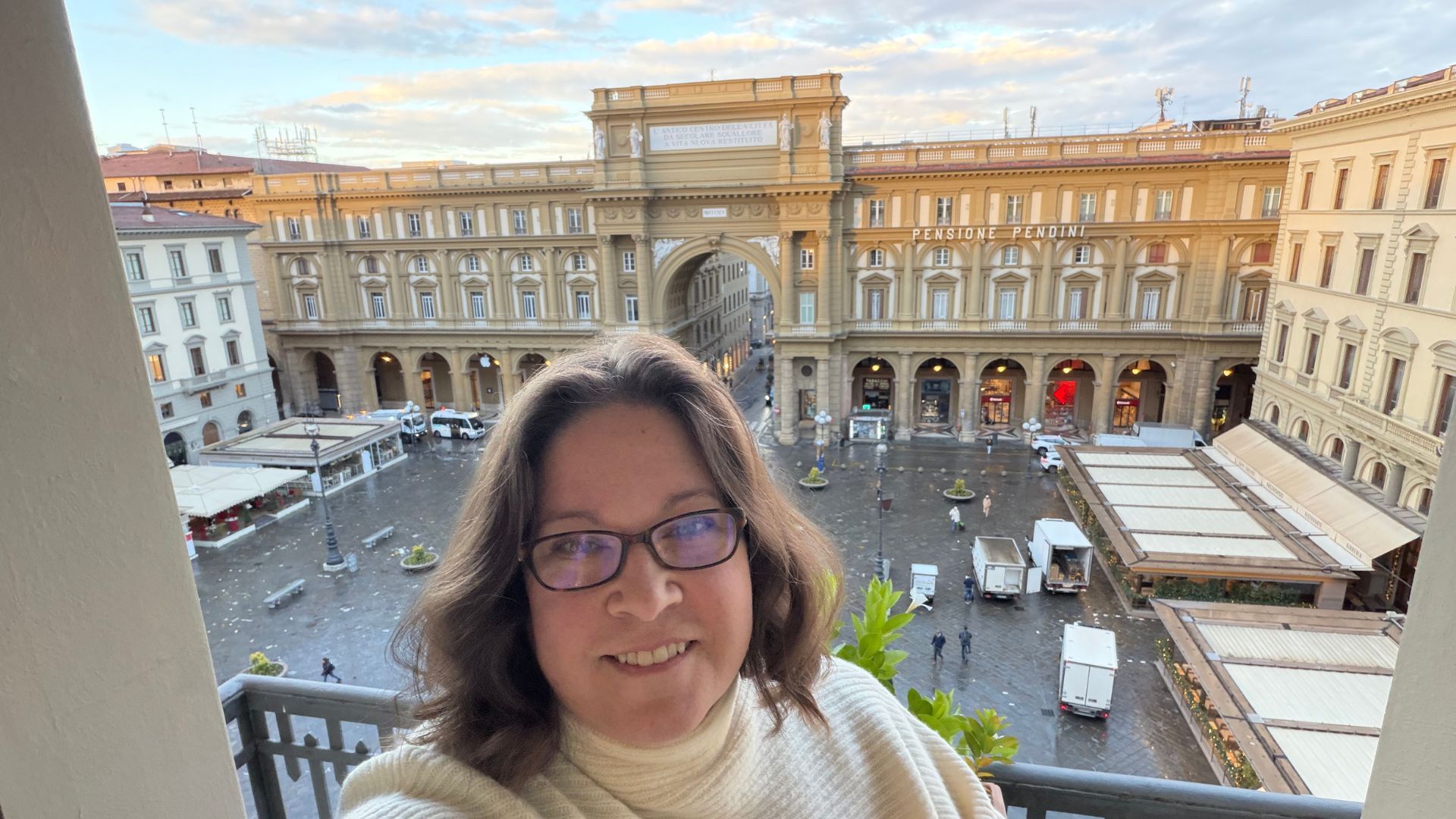 A woman smiles for a selfie from a balcony overlooking Florence’s Piazza della Repubblica. The large historic arch and surrounding elegant buildings frame the busy square below, where delivery trucks and pedestrians move across the wet cobblestone plaza at sunrise.