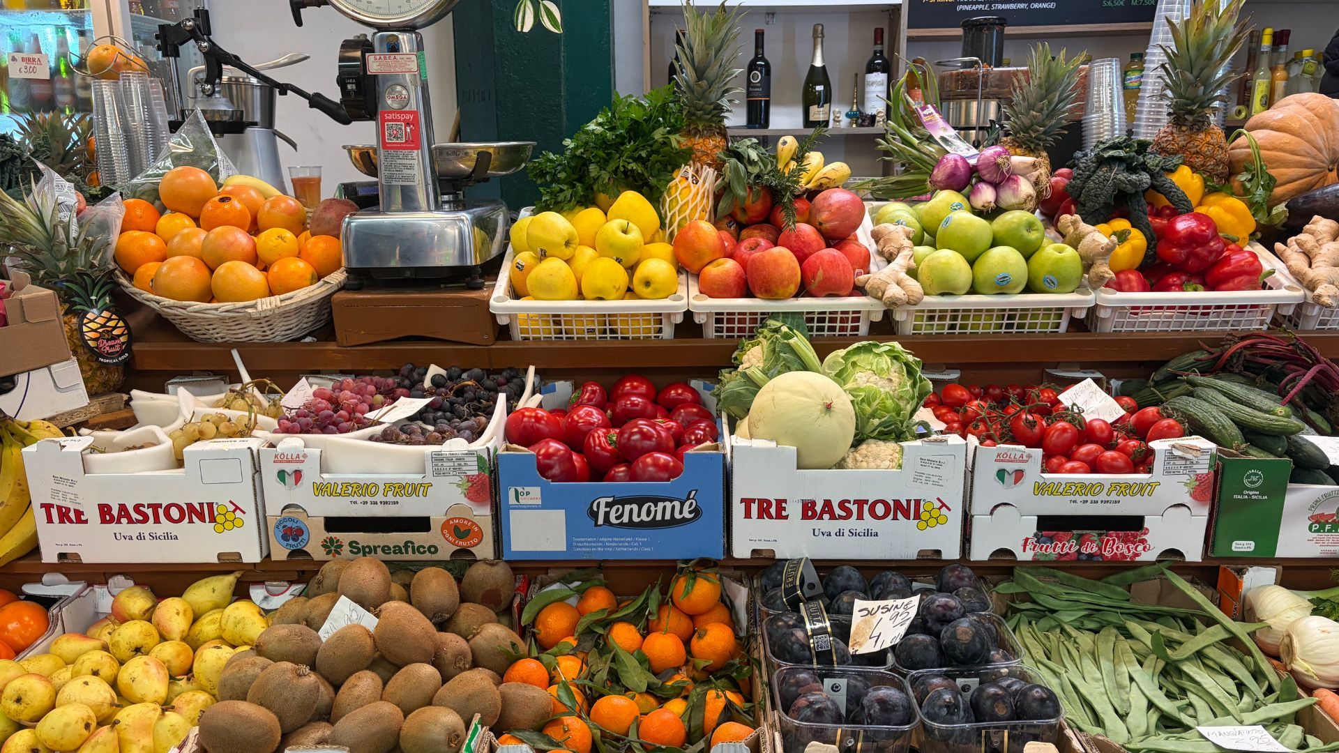 A colorful fruit and vegetable stall displays oranges, lemons, apples, peppers, kiwis, grapes, melons, zucchini, and other fresh produce in cardboard crates. A scale, juicer, and bottles of wine are visible in the background of the busy market stand.