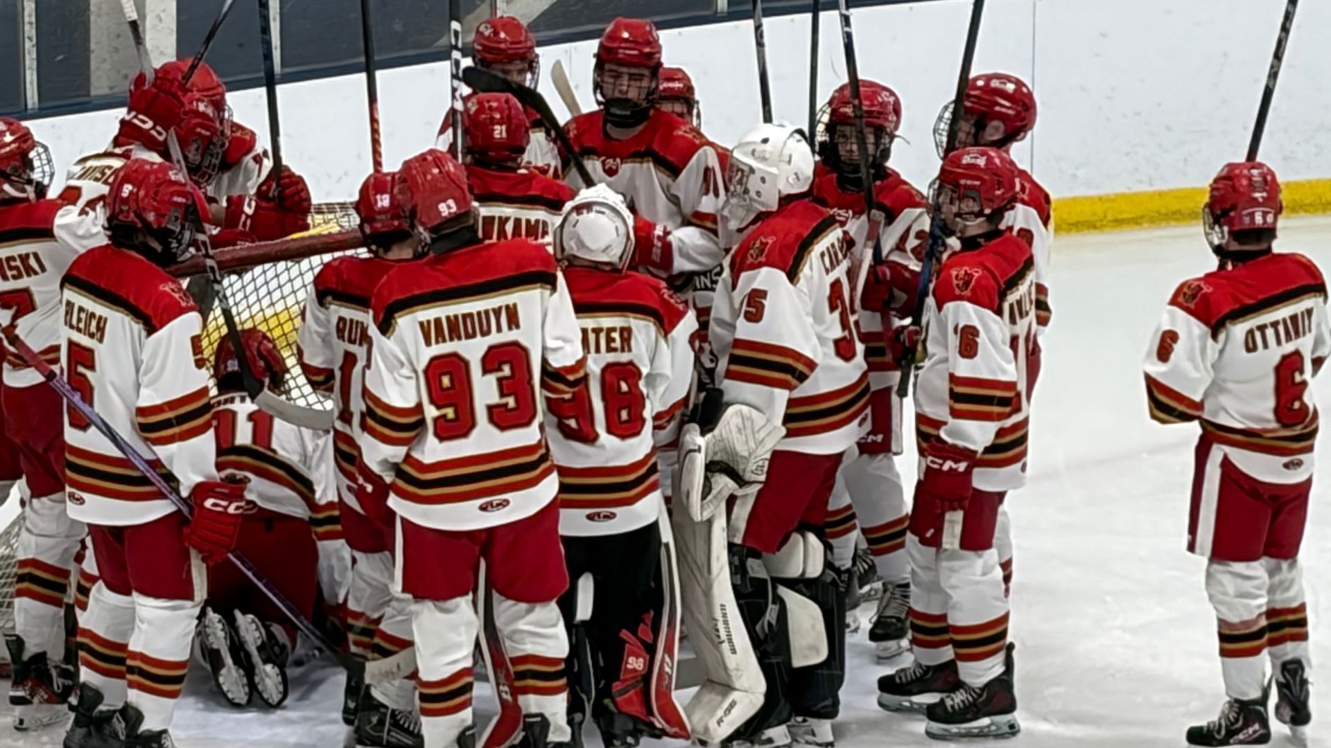 A youth ice hockey team in red and white uniforms gathers tightly around the net before a game, raising their sticks and listening to each other. The players’ jerseys display various numbers and names as they stand on the ice near the boards.