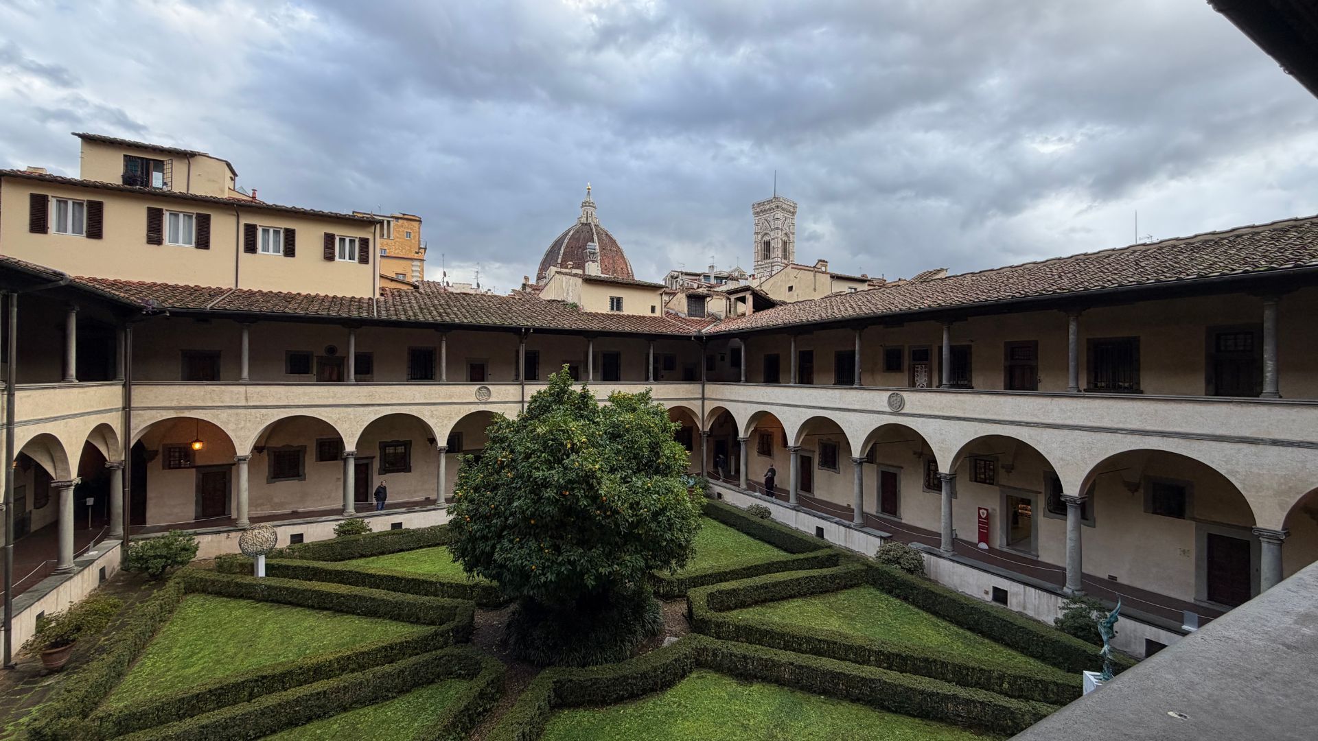 A quiet rectangular cloister garden with manicured hedges and a central citrus tree is surrounded by an arched walkway and two-story colonnade. Florence rooftops, Brunelleschi’s dome, and Giotto’s bell tower rise in the distance under a cloudy sky.