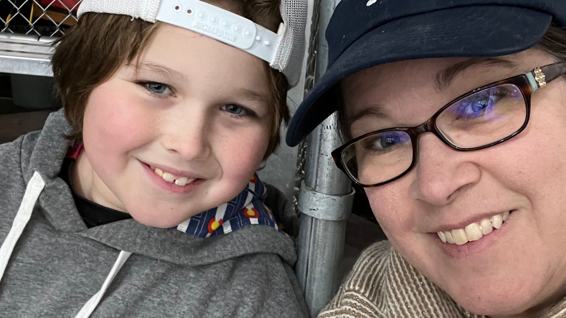A smiling woman and her young son take a close-up selfie at an indoor hockey rink. The boy wears a backwards white cap and a gray hoodie, while the woman wears glasses and a navy cap. Metal railings and rink equipment are visible behind them.