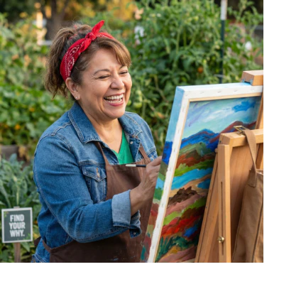 A woman in her 60s stands outdoors painting on an easel, smiling as she works. The scene reflects creativity, joy, and the confidence that comes from finding your why.