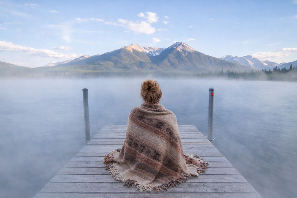 Shawl draped woman seated on a jetty overlooking a mountain lake.