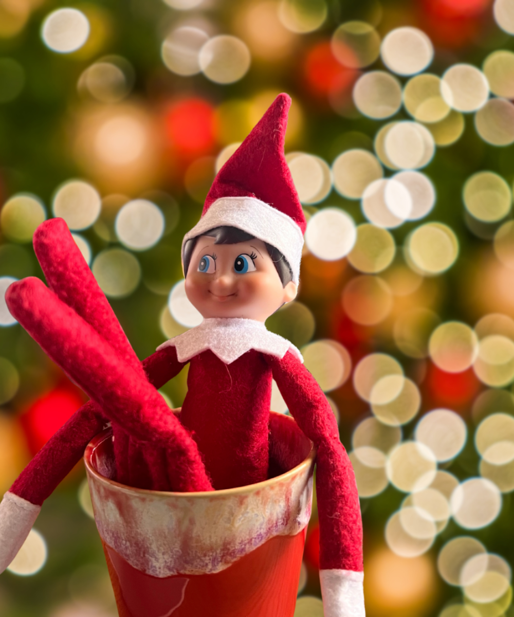A red and white elf doll sits in a red cup with a blurred background of festive lights.