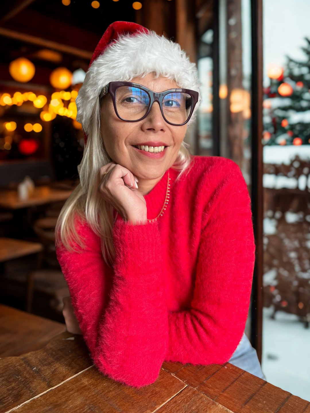 Andrea wearing a red sweater, Santa hat, and glasses smiles while sitting at a wooden table in a festive indoor setting with holiday lights.