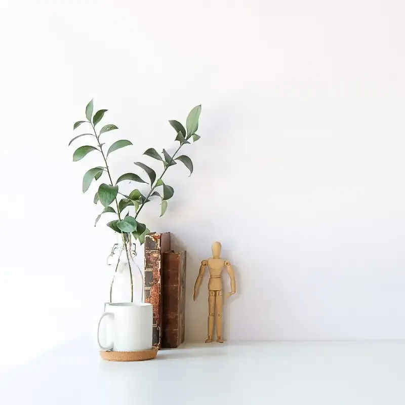 white greenery in white vase next to white coffee cup old book and wooden human form model