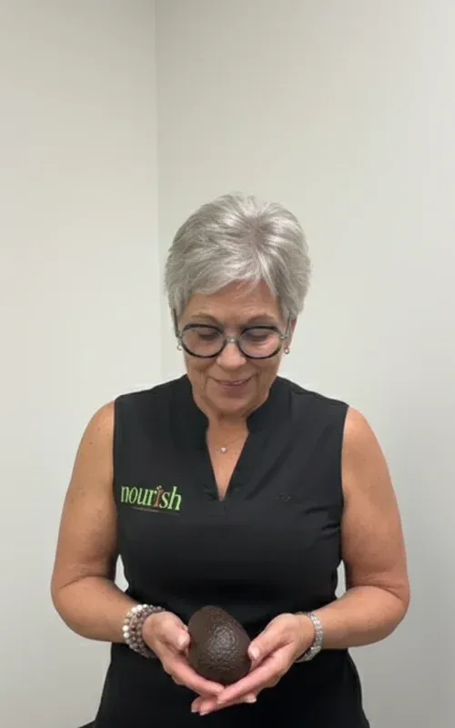 Woman over 50 smiling while holding a ripe avocado indoors, representing real food, nourishment, and the connection between avocados and better sleep for sustainable weight loss.